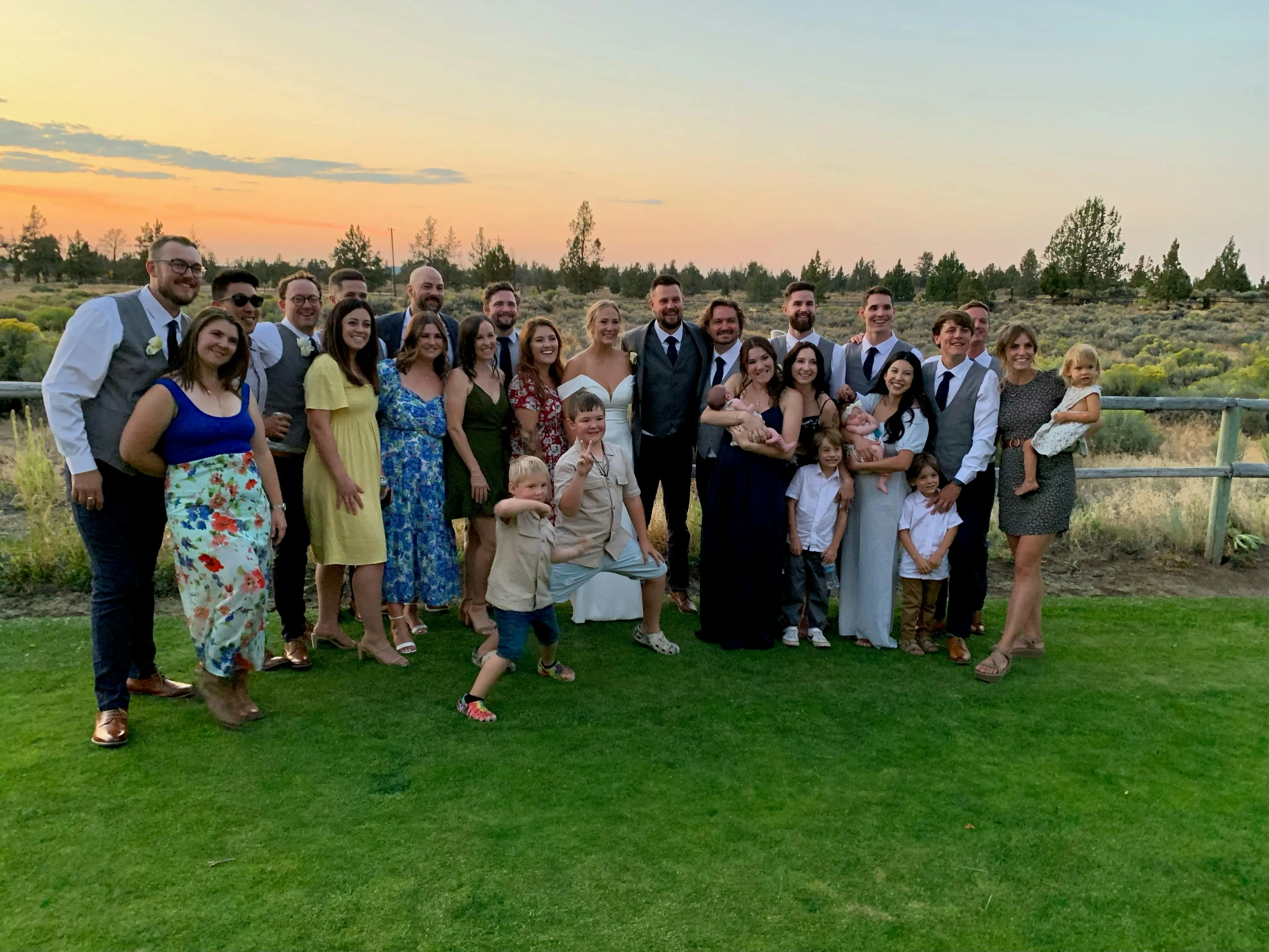 Large group of wedding guests, including the bride in a white dress and groom, kids and families, posing together on a grassy lawn at sunset with high desert scrub and pines behind them